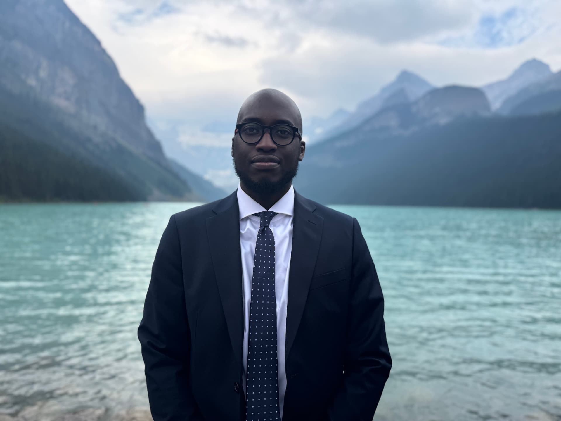 Andrew Adelowokan, founder of The Resilient Institution, photographed in a navy suit before a glacial lake and mountain peaks